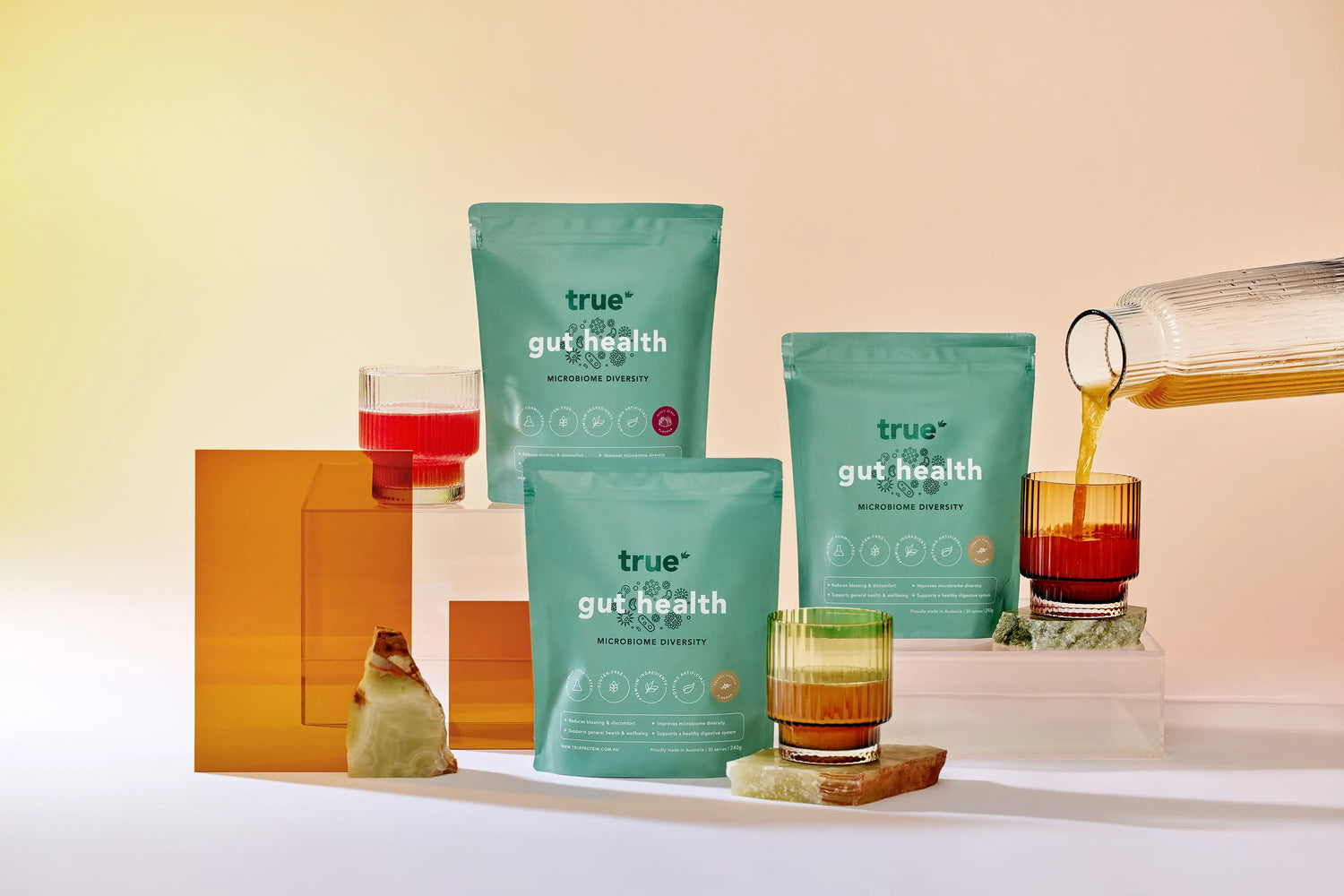 True Protein "True Gut Health" probiotic powder pouches (three teal resealable bags) styled on a pastel studio backdrop with ribbed drinking glasses, amber acrylic blocks and a pitcher pouring a drink — gut health supplement product shot for ecommerce and SEO.