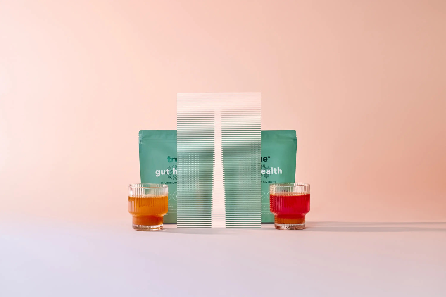 True Protein "Gut Health" supplement pouch with two glasses of orange and red probiotic drinks on a pastel pink background — gut health powder product shot, probiotic/prebiotic supplement.