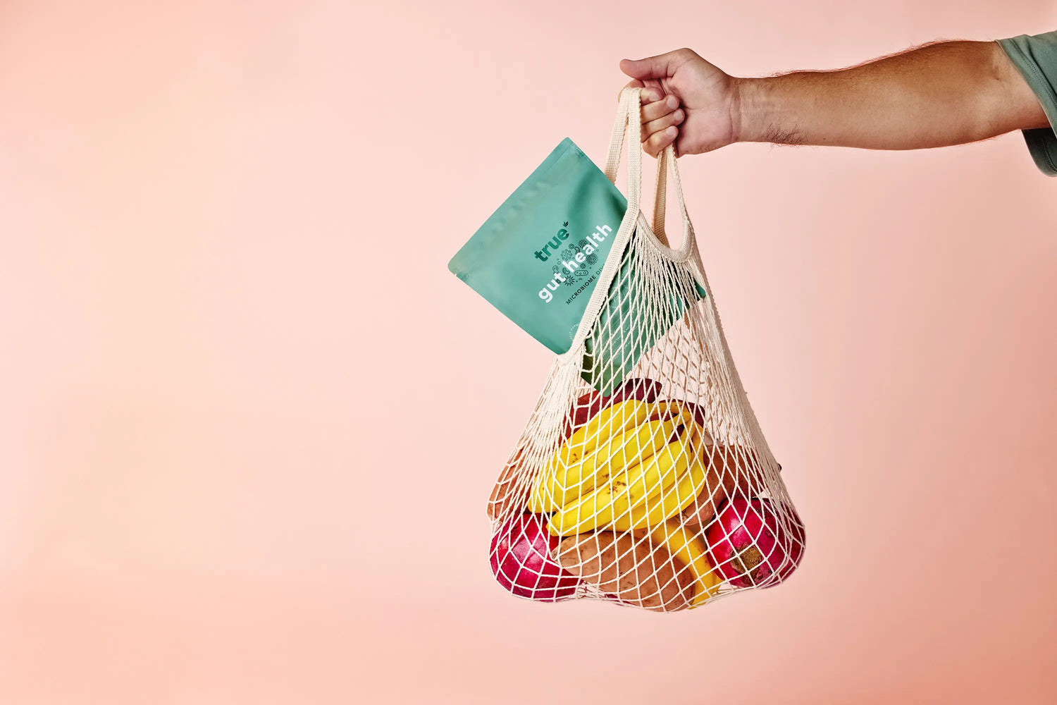 Hand holding reusable mesh grocery bag filled with bananas, apples and kiwi plus a teal "True gut+health" probiotic powder pouch against a soft pink background