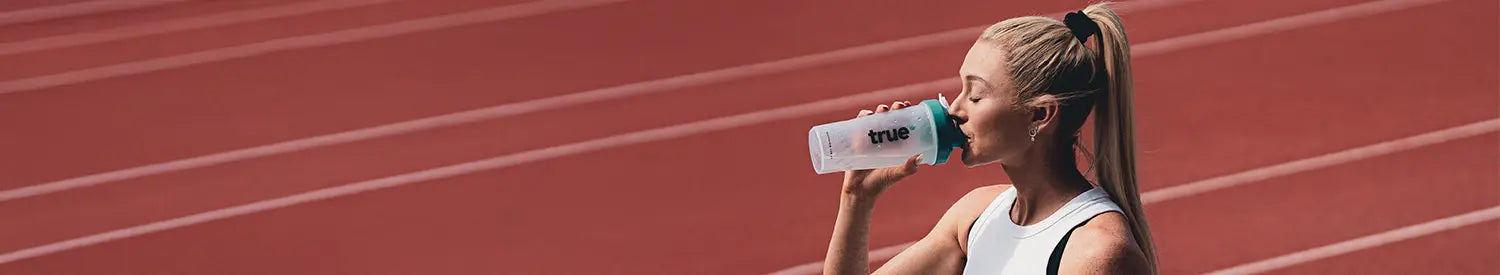 Female athlete on a red outdoor running track drinking from a "True" branded shaker bottle during training session
