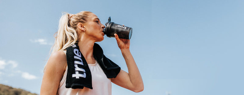 Female athlete with ponytail drinking from a black protein shaker bottle and wearing a black "true" towel over her shoulders during an outdoor sunny workout, sports hydration and fitness concept