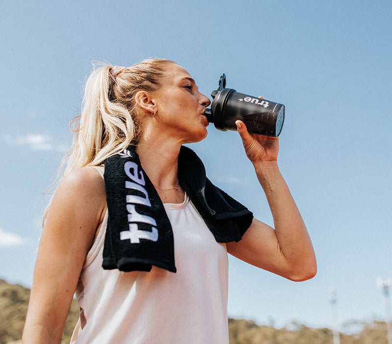 Blonde female athlete outdoors drinking from a black "True" shaker bottle with a black "True" towel over her shoulders, wearing a white tank top against a clear blue sky after a workout