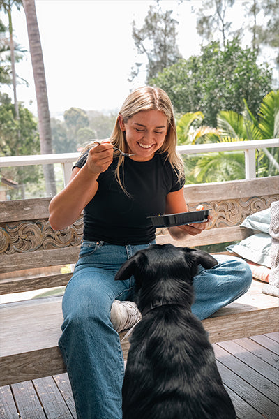 Young woman sitting on a sunlit wooden deck bench eating a prepared meal from a black container while smiling at a black dog begging for food, with palm trees and lush greenery in the background.