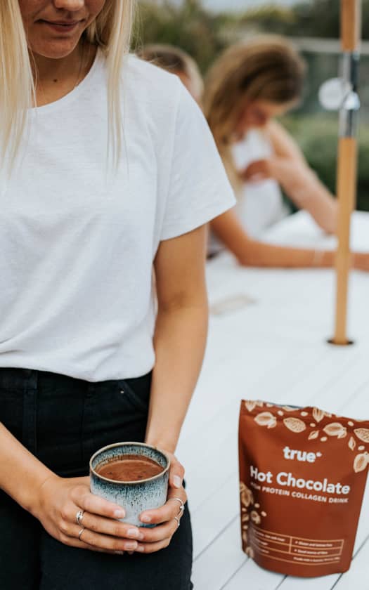 Woman holding a ceramic mug of hot chocolate beside a True Hot Chocolate protein collagen drink pouch on a white outdoor patio table