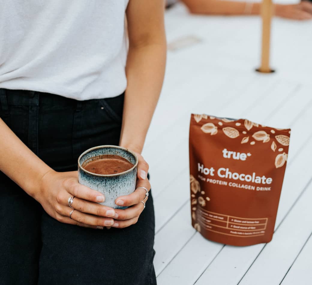 Person holding a ceramic mug of True Hot Chocolate collagen protein drink next to a brown True Hot Chocolate pouch on a white wooden deck, cozy beverage product shot.