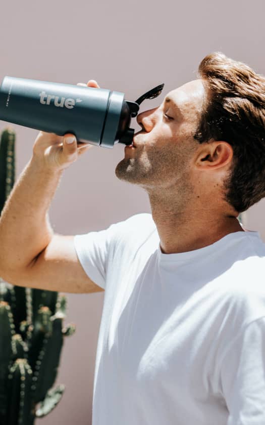 Man in white T-shirt drinking from a blue "true" insulated water bottle outdoors next to a cactus, summer sunlight, lifestyle hydration product photo for active adults.
