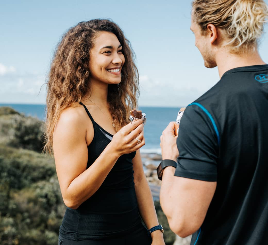 Young athletic couple in black activewear smiling and eating energy bars on a rocky coastal trail with ocean in the background.