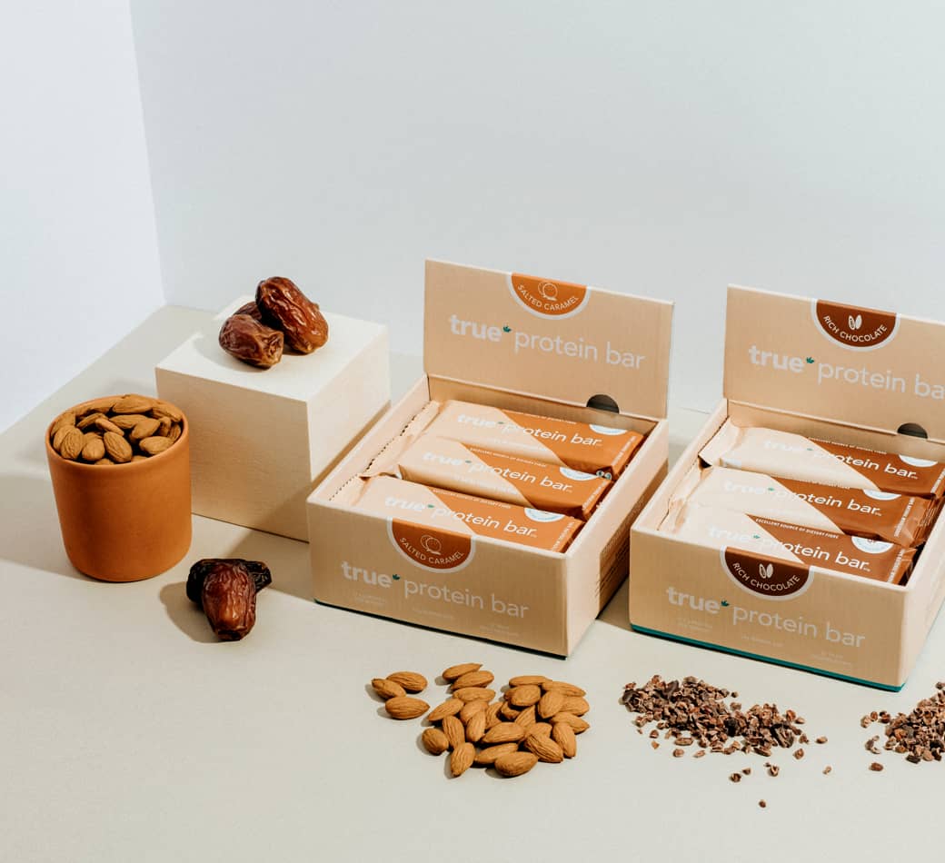 Boxes of True protein bars (salted caramel and rich chocolate) displayed with almonds, whole dates and cacao nibs on a neutral tabletop — ecommerce product shot for health snack bars.