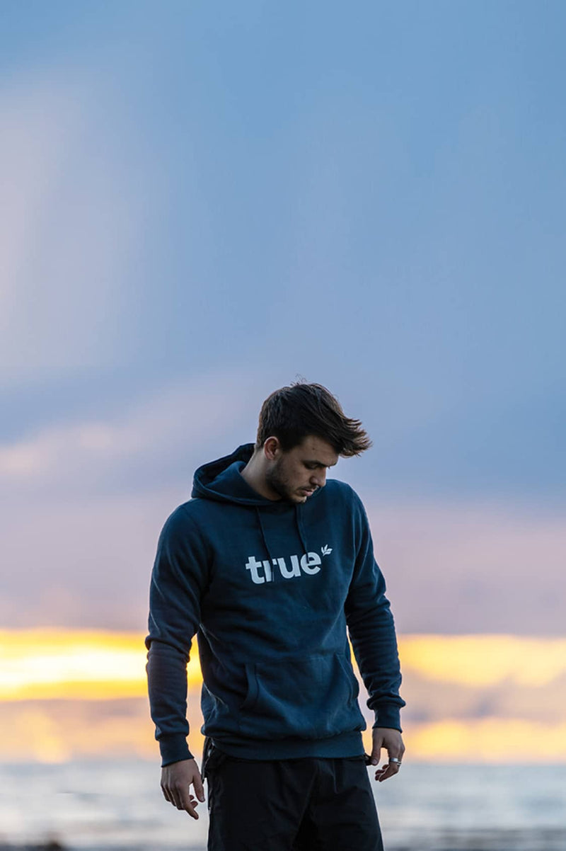 Man wearing navy "true" logo hoodie standing on a beach at sunset — men's casual streetwear hoodie product photo, mobile banner for coastal lifestyle and outdoor apparel.