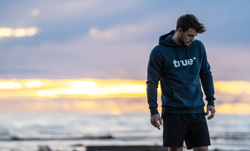 Man wearing navy "true" hoodie and black shorts standing on a rocky beach at sunset, lifestyle apparel hoodie photographed on ocean coastline with warm sky background