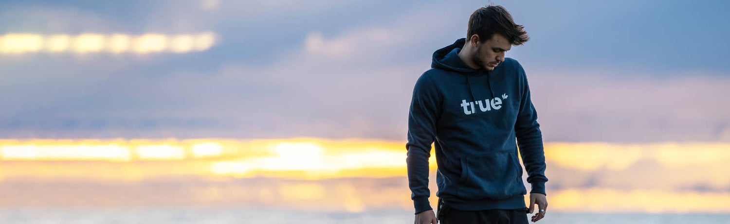 Young man wearing navy "true" hoodie standing on a coastal beach at sunset with blurred ocean and golden sky — men's lifestyle apparel hoodie product image