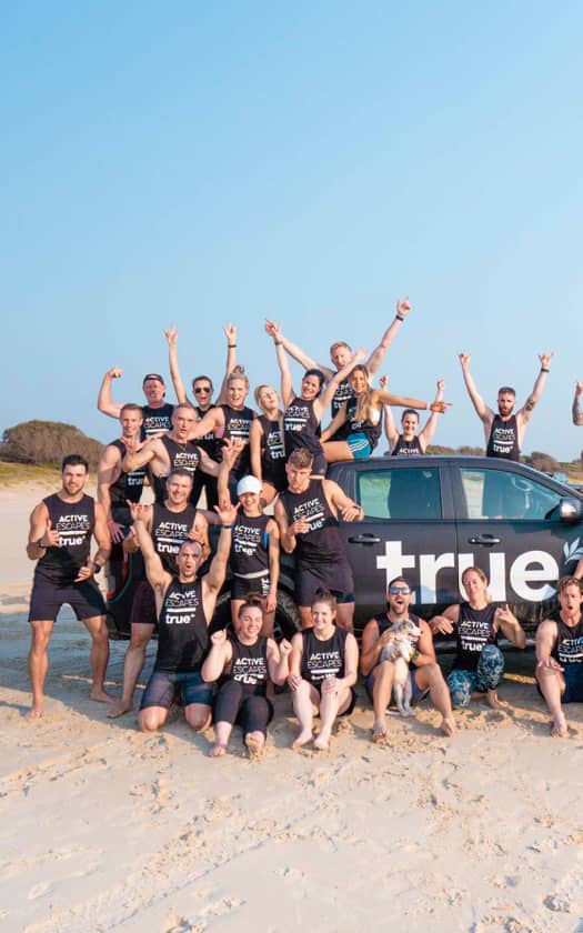 Group of fit adults in matching "Active Escapes" tank tops posing on a sandy coastal beach around a branded "True" vehicle, smiling and raising hands — beach fitness retreat, group training and team wellness event on a sunny day.