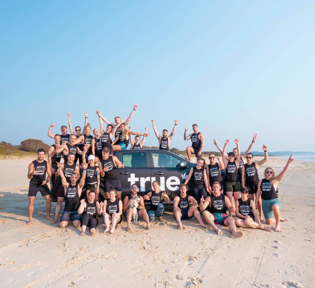 Large group of ~30 young adults wearing "True" and "Active Escapes" shirts posing around a branded "true" vehicle on a sunny sandy oceanfront beach, cheering with arms raised — coastal team/community retreat image.