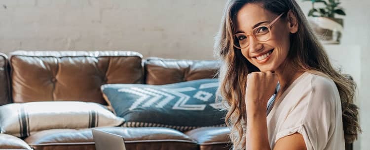 Young woman working on a laptop in a cozy modern living room with leather sofa and decorative pillows, remote work lifestyle scene