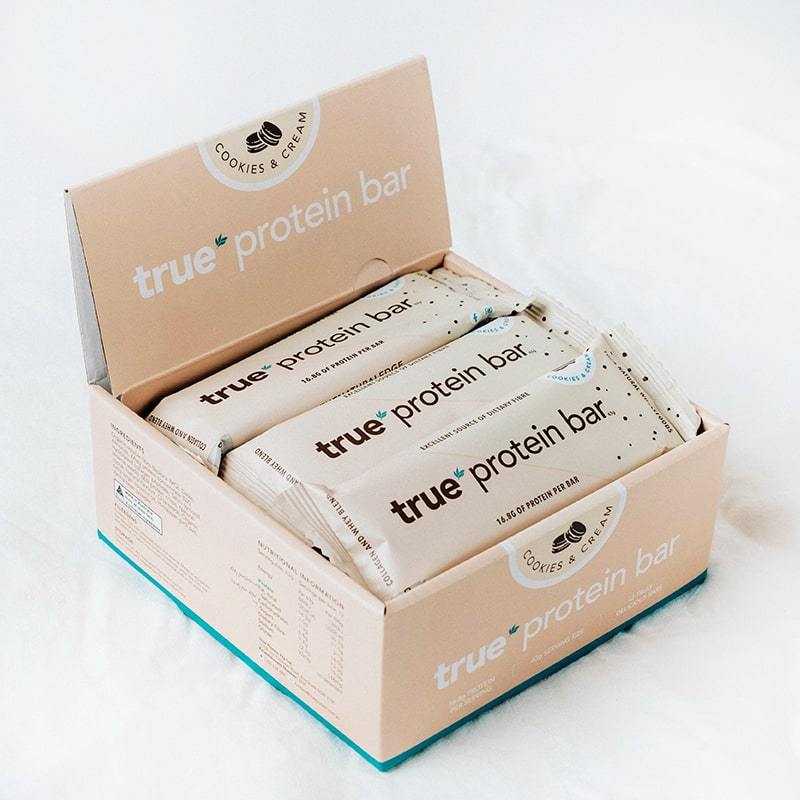 Box of True Protein Bar (Cookies &amp; Cream flavor) with multiple individually wrapped beige bars displayed in an open retail carton on a white surface.