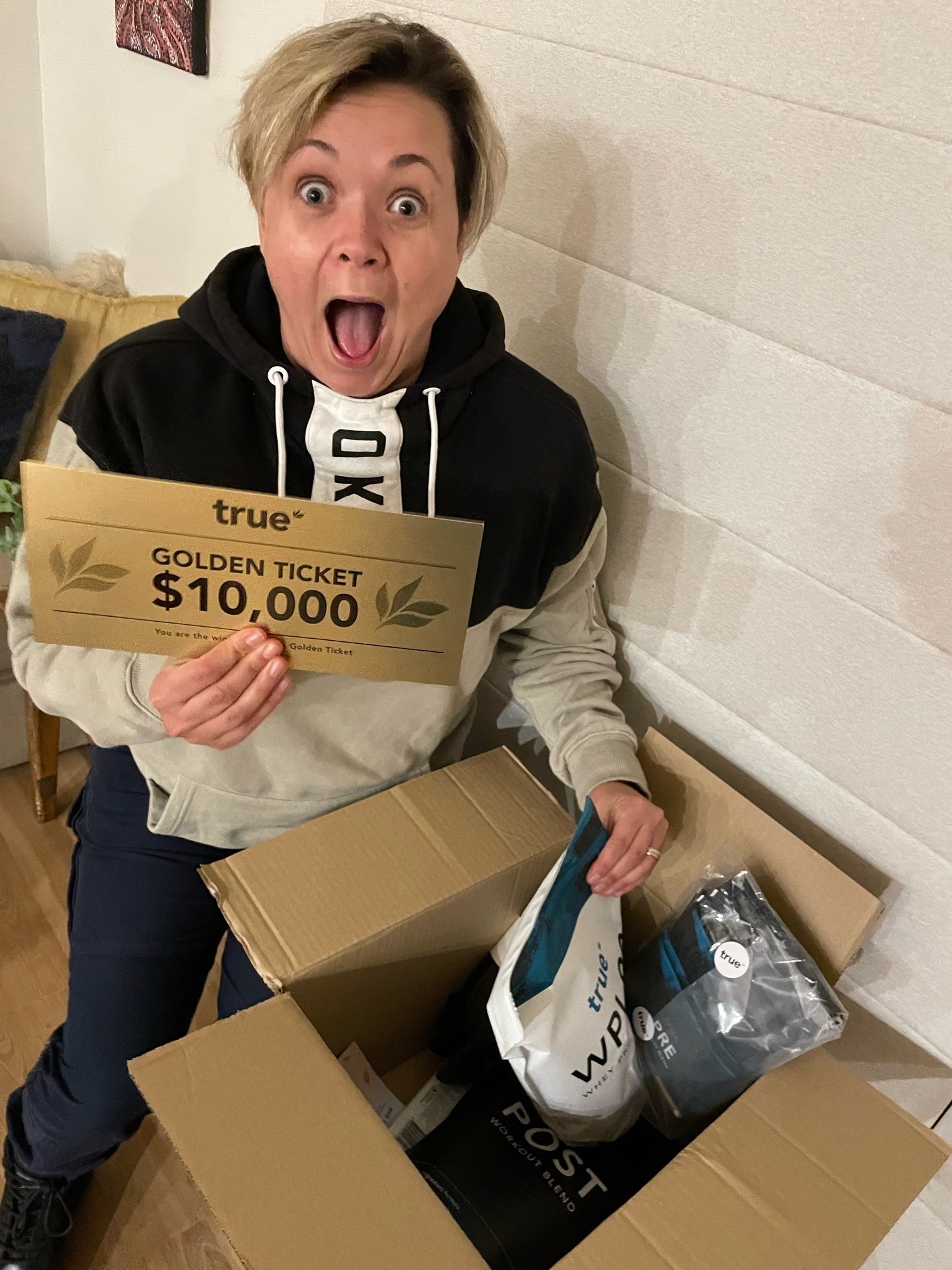 Person with surprised expression holding a gold "true" Golden Ticket reading "$10,000" while unboxing True nutrition products from an open cardboard box — unboxing True protein and workout supplement bundle.