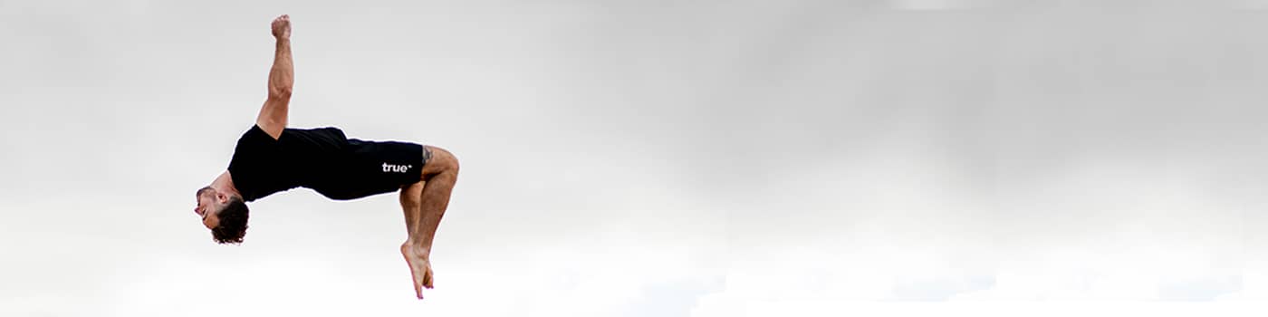Athlete in black workout clothes performing a mid-air backflip outdoors against a cloudy sky — fitness acrobatics banner for workout, training, and healthy lifestyle.