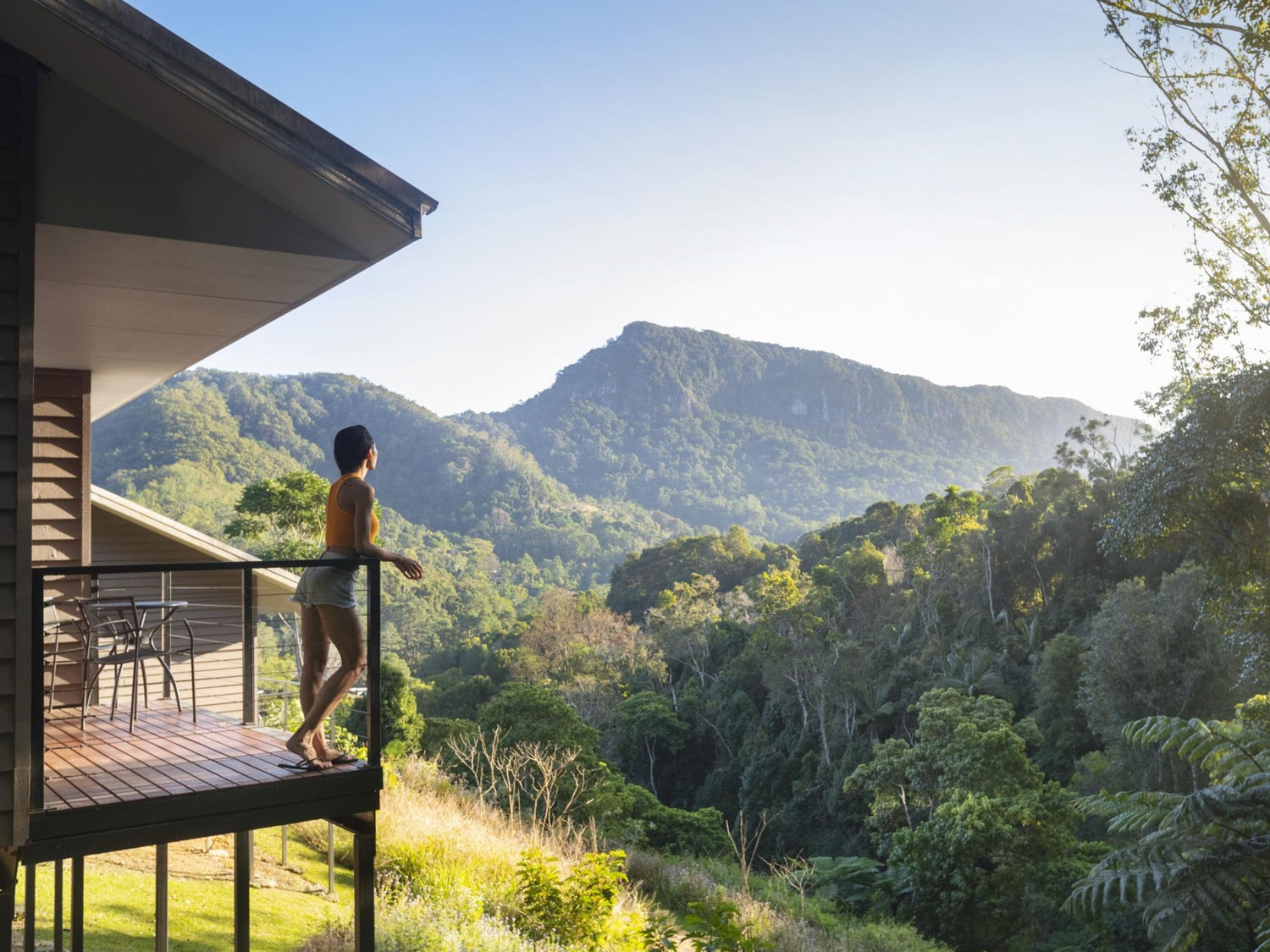 Woman on a wooden balcony of an eco-retreat cabin overlooking a lush subtropical rainforest valley and mountain ridge at sunrise — wellness retreat, nature view, scenic mountain landscape.