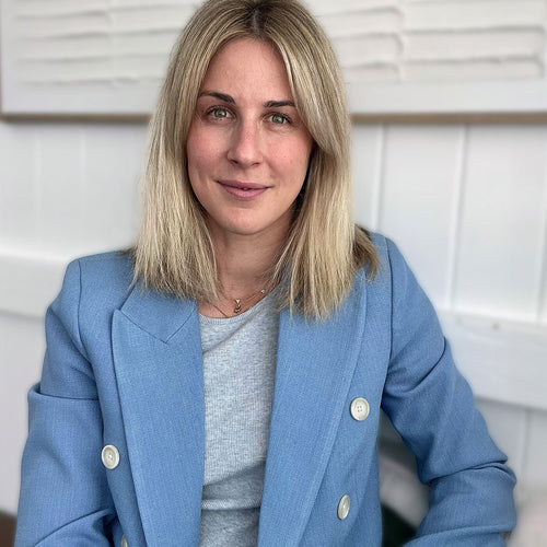 Professional headshot portrait of a blonde woman wearing a light blue double-breasted blazer and grey top, seated against a white paneled interior background — business portrait, corporate headshot, lifestyle branding image.