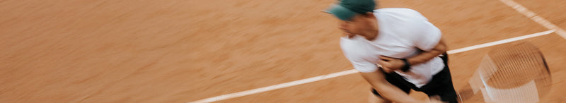 Blurred action shot of a male tennis player in a white shirt and green cap hitting a forehand on an outdoor clay tennis court, motion-blurred racket swing and court lines visible.