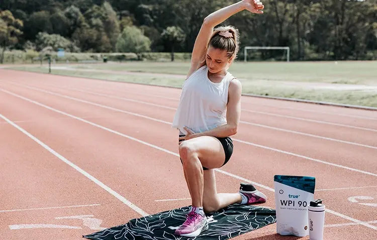 Woman athlete stretching on an outdoor running track next to a towel, pink running shoes, True WPI 90 whey protein pouch and shaker bottle — outdoor fitness workout scene at an athletics track.