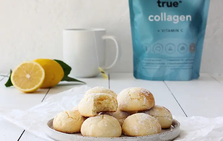 Plate of lemon crinkle cookies dusted with powdered sugar on a white table with a sliced lemon, white mug, and a blue True Collagen pouch in the background