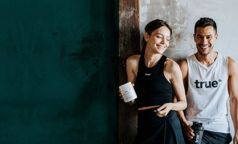 Two smiling fit adults in True-branded activewear holding a True supplement tub and shaker bottle, posing against a rustic textured wall — fitness supplements, protein powder, workout nutrition.