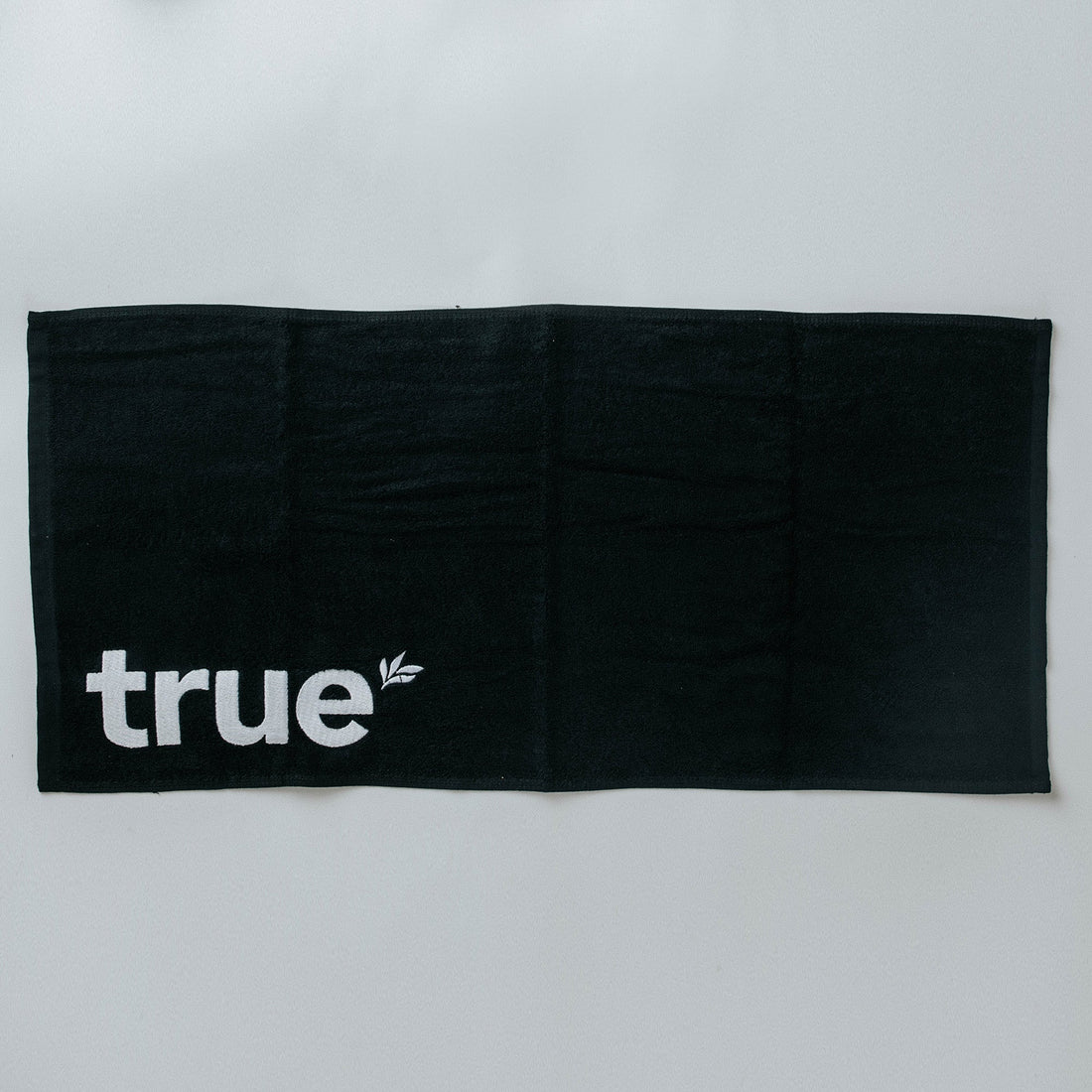 Black cotton gym towel laid flat with white embroidered "true" logo in lower left corner — workout towel for gym, crossfit, yoga and fitness use.