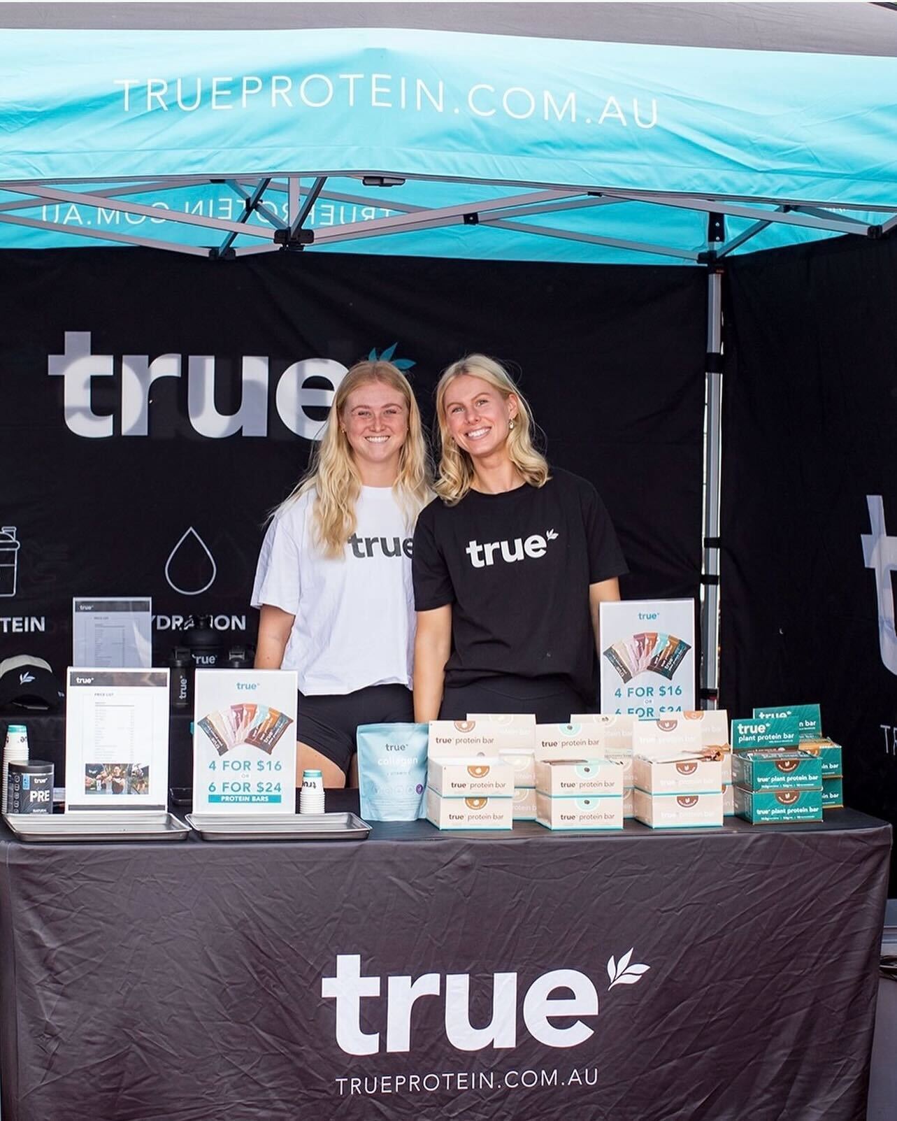 Two smiling female staff wearing "true" shirts standing behind a True Protein market stall displaying protein bars, supplements and signage under a turquoise TrueProtein.com.au canopy — Australian outdoor health food market.