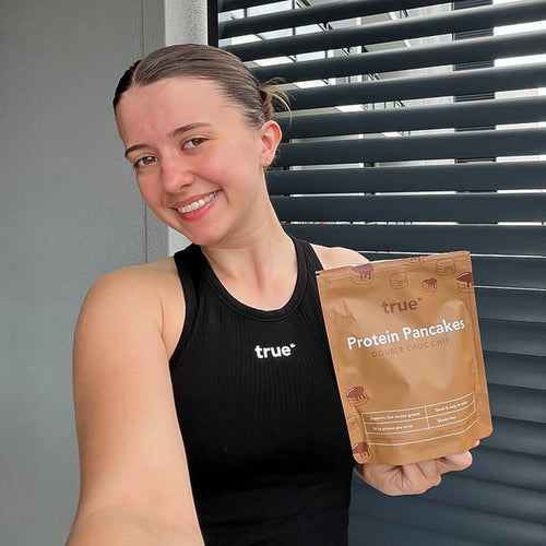 Smiling young woman in black "true" tank top holding a pouch of True Protein Pancakes Double Choc Chip on an apartment balcony with louvered blinds — fitness-focused, high-protein pancake mix product image.