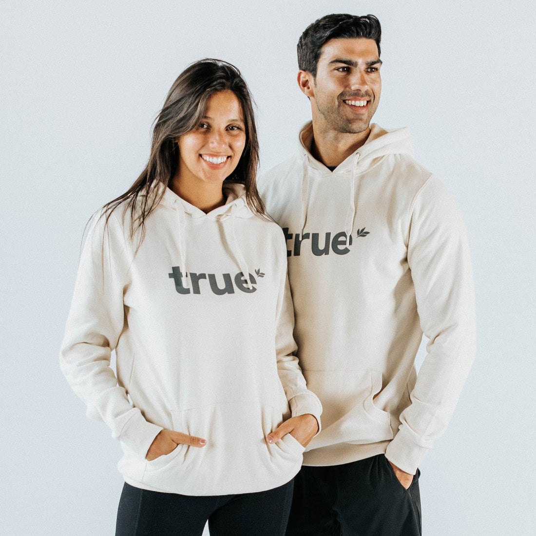 Two models wearing cream unisex hoodies with "true" logo across the chest, drawstring hood and front kangaroo pocket — studio lifestyle shot for cream hoodie product listing.