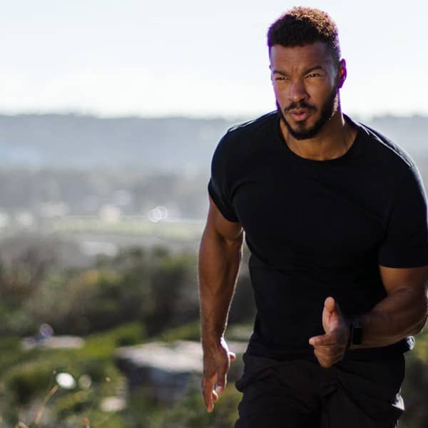 Athletic man in a black performance t-shirt and smartwatch sprinting uphill on a scenic outdoor trail with a blurred cityscape in the background — outdoor running fitness apparel product image.