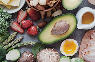 Keto diet ingredients on dark slate: avocado halves, soft‑boiled egg, bacon, asparagus, broccoli, mushrooms, strawberries, spinach and olive oil — low‑carb healthy meal inspiration