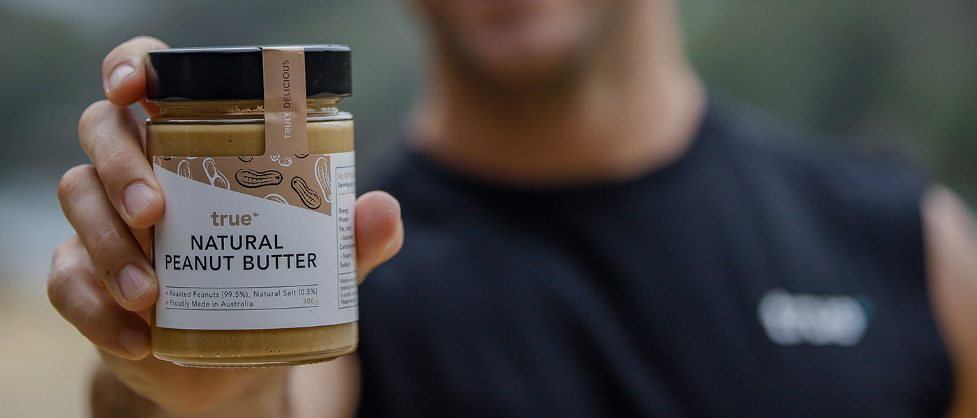 Person holding a jar of True Natural Peanut Butter (300g) with visible label "Proudly Made in Australia" — natural peanut butter jar, healthy keto-friendly spread, Australian-made product