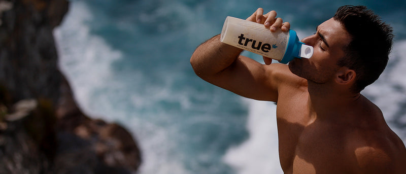 Shirtless man drinking TRUE keto protein shake from branded shaker bottle on an Australian coastal cliff with ocean waves in background