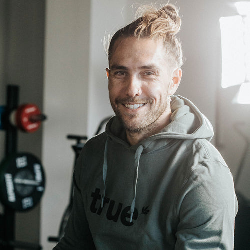 Smiling man with top knot wearing a green "true" hoodie seated in a bright indoor gym, with weight plates and barbell equipment in the blurred background.
