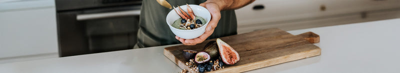 Hands holding a healthy meal-prep breakfast bowl with green smoothie, granola, blueberries and figs on a wooden cutting board on a modern home kitchen countertop.