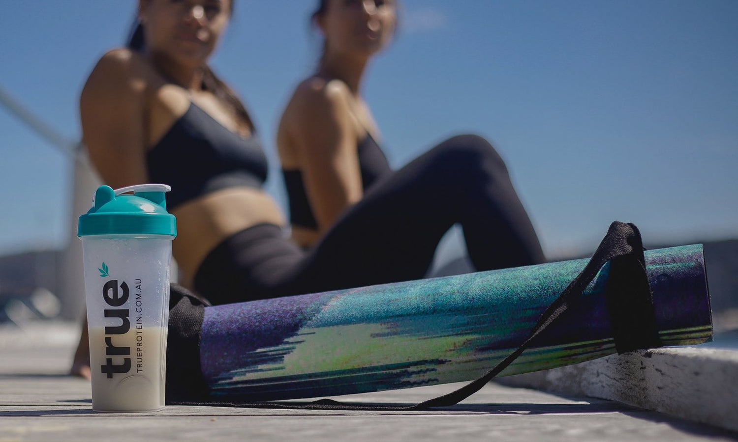Protein shaker bottle labeled "true" next to colorful yoga mat on a wooden dock with two blurred women in black activewear doing an outdoor workout by a sunny marina under a clear blue sky.