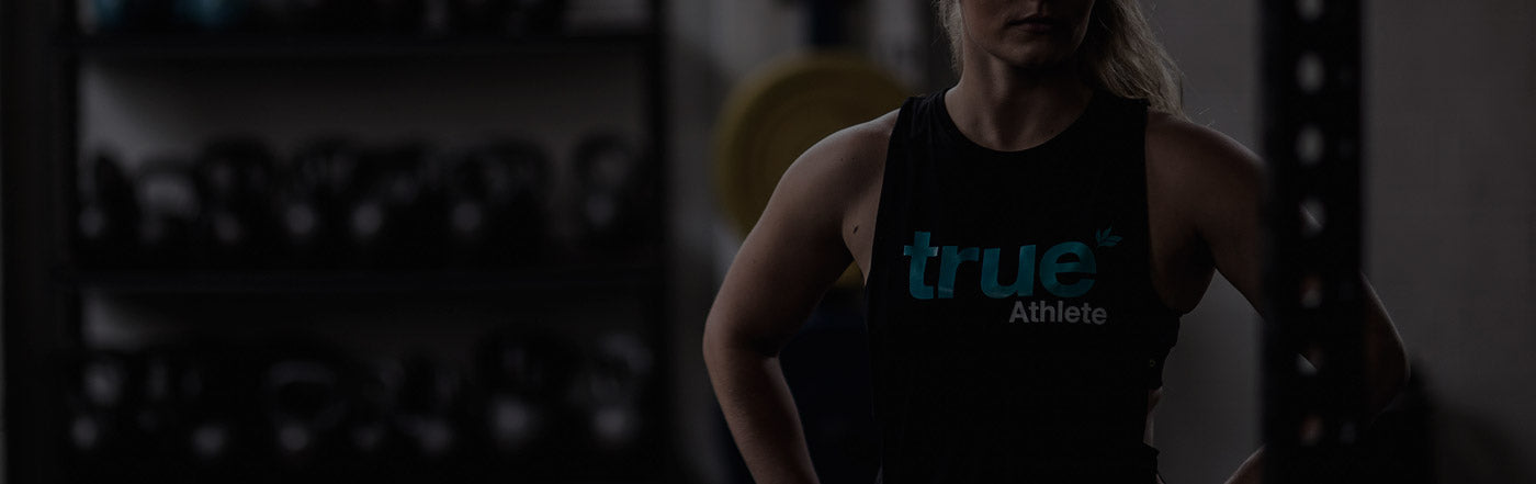 Female athlete wearing a "true Athlete" tank top standing in a dim CrossFit-style gym with kettlebells on shelves and weight plates in the background