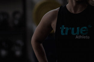 Person wearing a black "True Athlete" sleeveless tank top in a dimly lit gym, showcasing fitness apparel and strength training atmosphere.