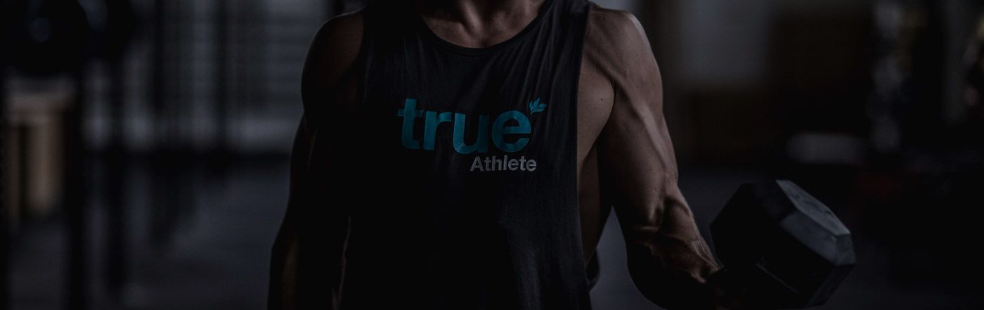 Muscular man wearing "true Athlete" tank top lifting a heavy dumbbell in a dimly lit commercial gym, strength training and bodybuilding workout session, fitness lifestyle.