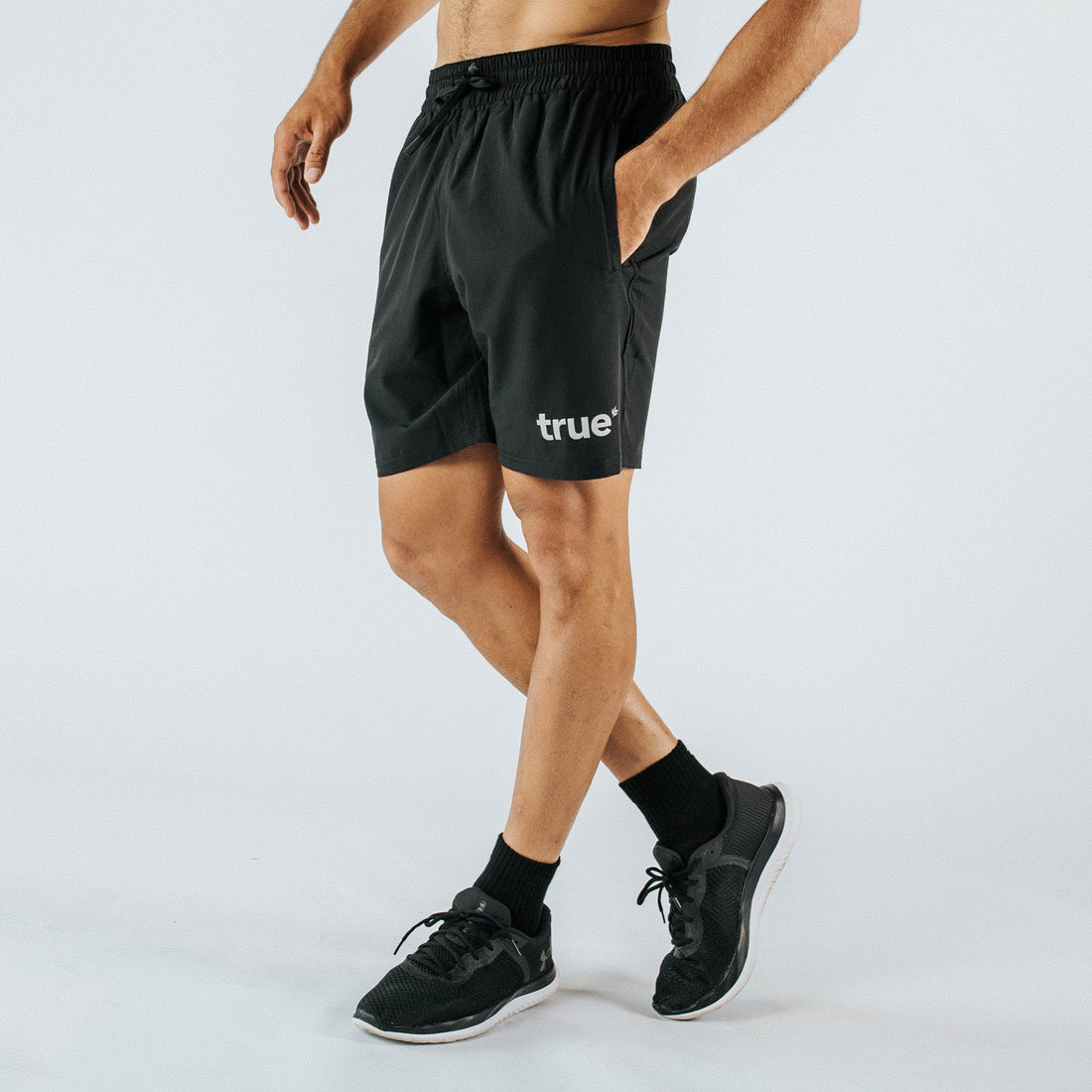Men’s black performance training shorts with elastic drawstring waistband, side pockets and white “true” logo, worn by athletic model for gym, running and workout — white studio background.