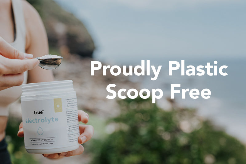 Woman scooping True Electrolyte powder from a white tub labeled "electrolyte" on a beach cliff background with large text "Proudly Plastic Scoop Free" — natural hydration supplement product photo for coastal lifestyle marketing.