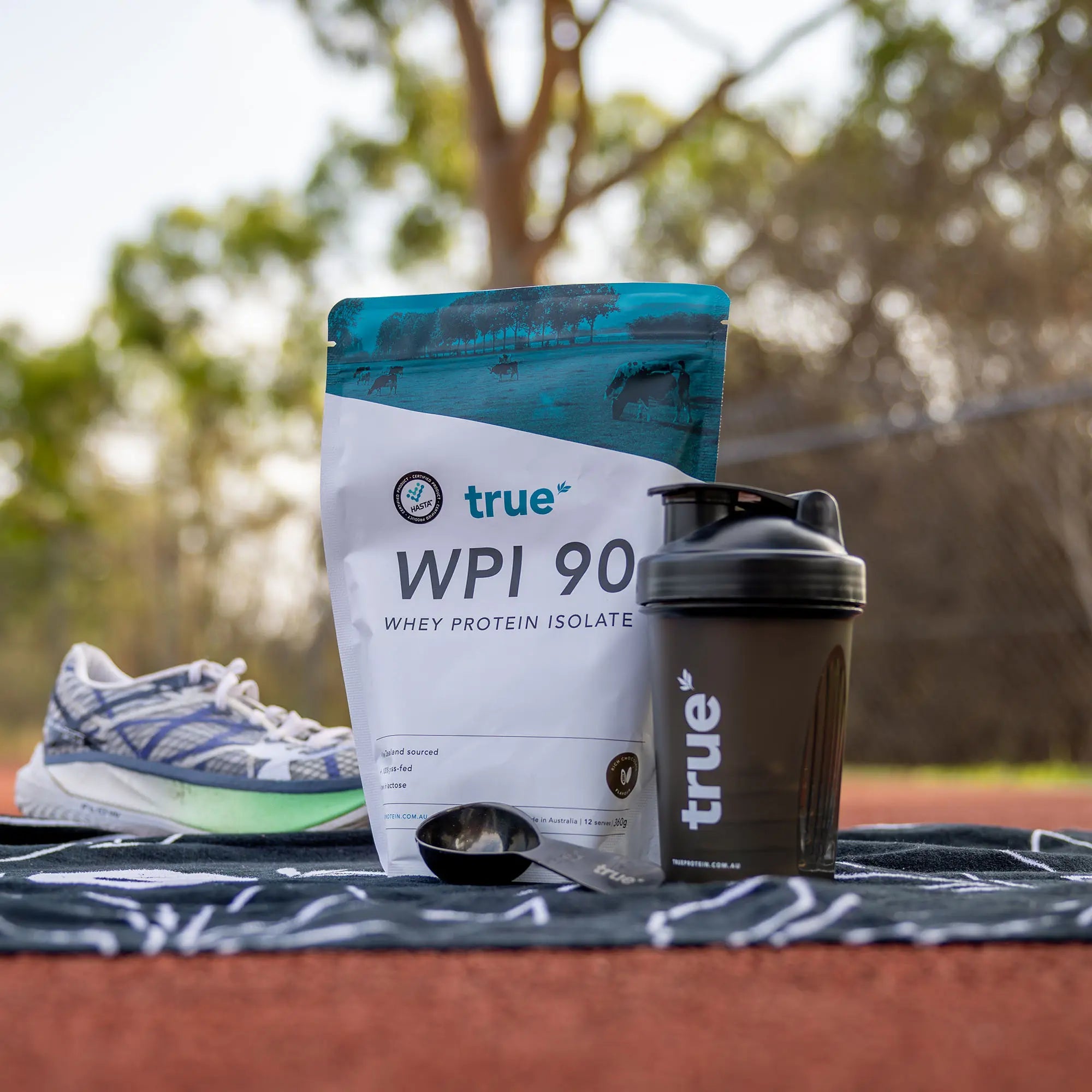 True WPI 90 whey protein isolate bag with True shaker bottle and scoop on a towel beside a running shoe on an outdoor track in Australia, fitness supplement and post-run recovery setup