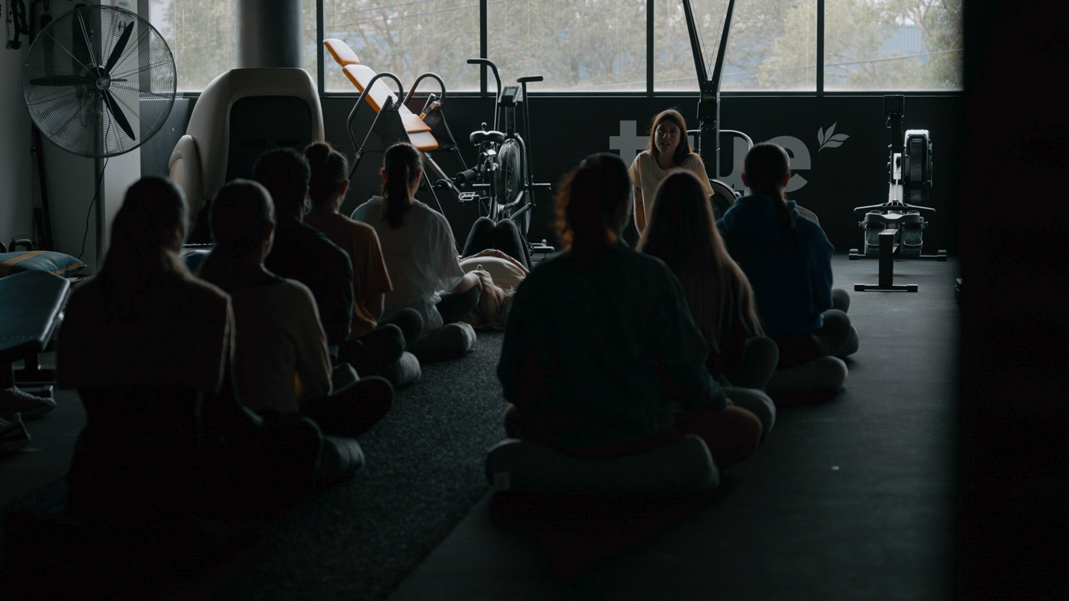 Group meditation at an R U OK? Day mental health workshop in a dim gym studio, instructor speaking to seated participants on cushions near exercise bikes and rowing machines.