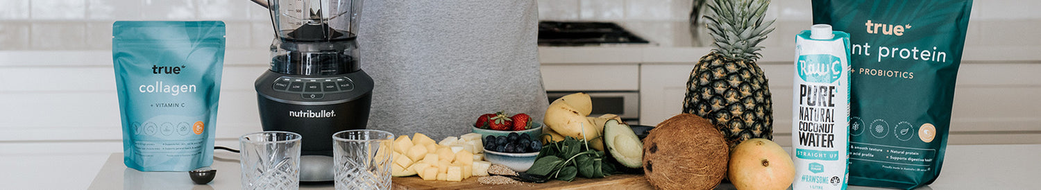 Kitchen counter with Nutribullet blender, True Collagen and True Plant Protein pouches, coconut water carton, pineapple, coconut, mango, banana, avocado, berries and spinach prepared for tropical smoothie recipes.