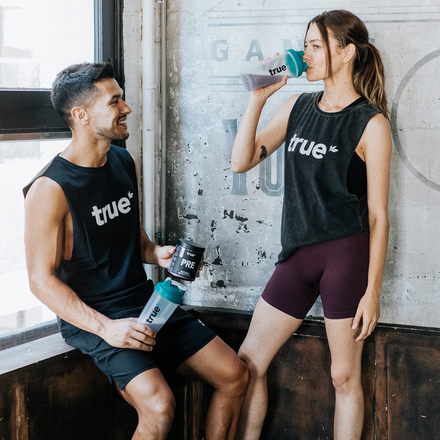 Two fit adults in an industrial gym wearing "true" logo tank tops, one sitting and holding a shaker bottle and pre-workout tub, the other standing drinking from a "true" shaker — fitness lifestyle, pre-workout supplement, gym apparel and shaker bottle product image.
