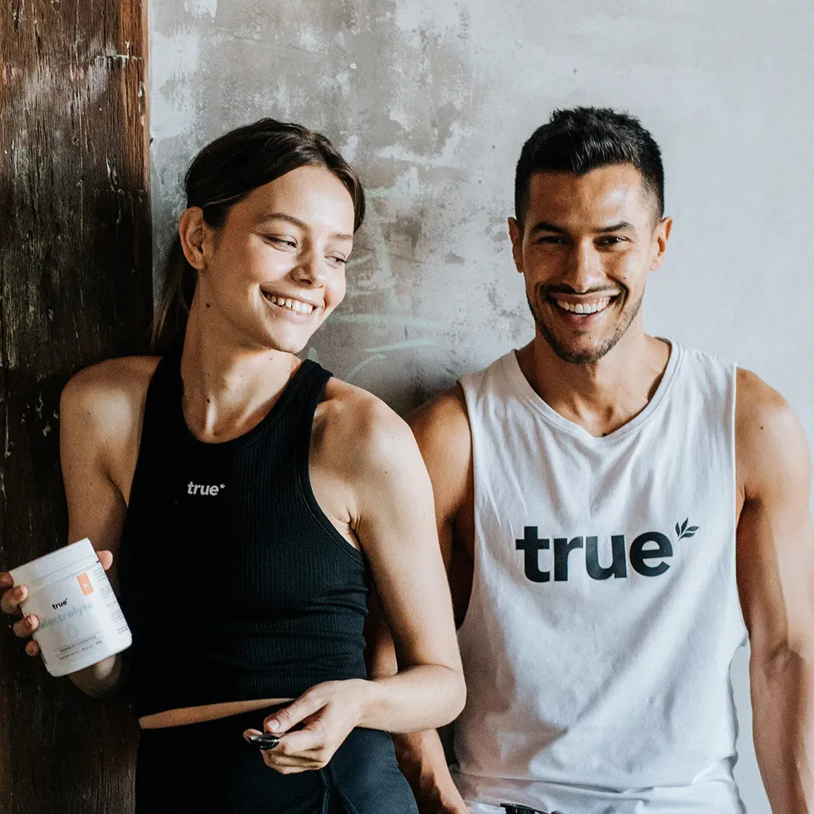 Young fit man and woman wearing "true" branded activewear, woman holding a supplement jar and spoon, both smiling against a textured concrete wall