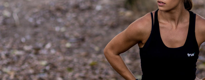 Woman wearing a black "true" branded women's tank top, cropped torso shot of a fit athlete with hands on hips on a woodland trail outdoor workout background.