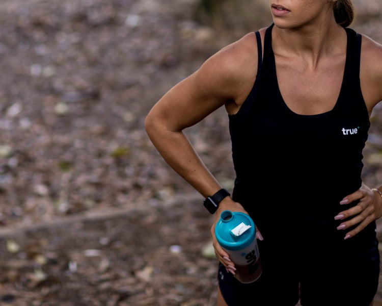 Athletic woman in black “true” tank top holding teal protein shaker bottle and wearing a smartwatch while pausing on a woodland trail